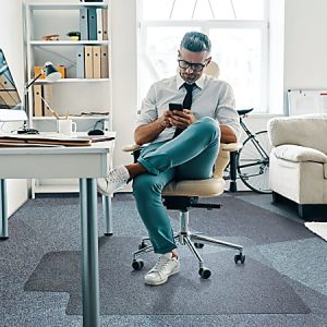 Man in office reading from phone, sitting on chair with legs crossed. Modern workspace with desk, lamp, shelves, and bicycle.