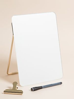 White dry-erase board on a stand with a pen and gold clip on a beige background.